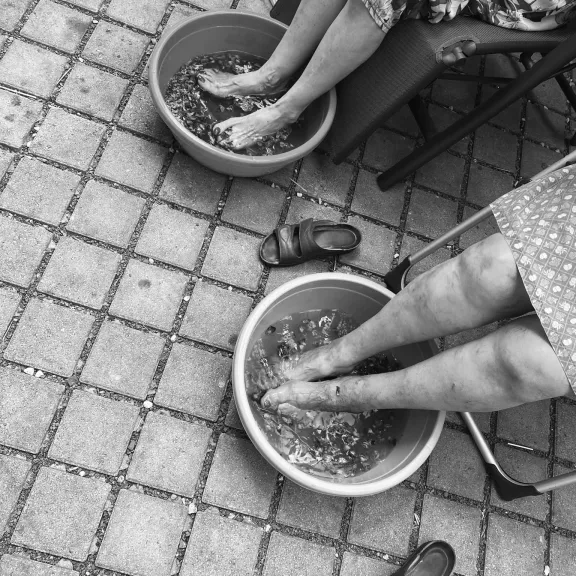 Twee personen genieten van een ontspannen voetbad in plastic bakken tijdens een pedicure, met slippers naast hen op de tegelvloer.