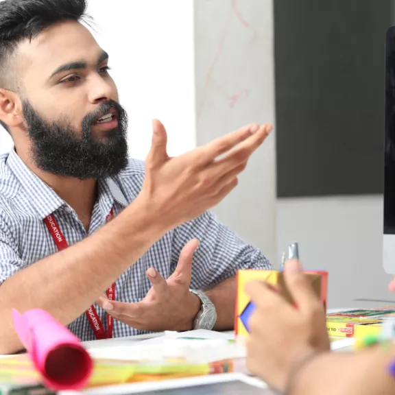 Een begeleidingsmoment waarbij een man uitleg geeft aan een andere persoon, met een betrokken en informatieve houding, zittend aan een bureau met diverse materialen en een computerscherm.