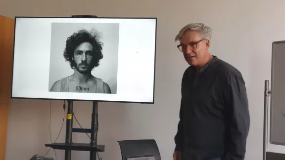 Een man geeft een presentatie in een leslokaal en staat naast een scherm waarop een zwart-wit portretfoto met de tekst '50mm' wordt getoond.