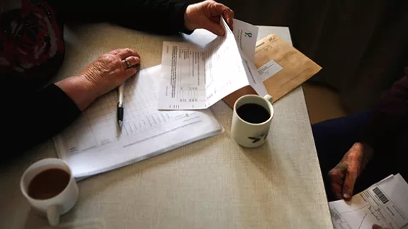 Twee mensen bespreken administratieve papieren aan een tafel, met kopjes koffie erbij en een pen klaarliggend op een document.